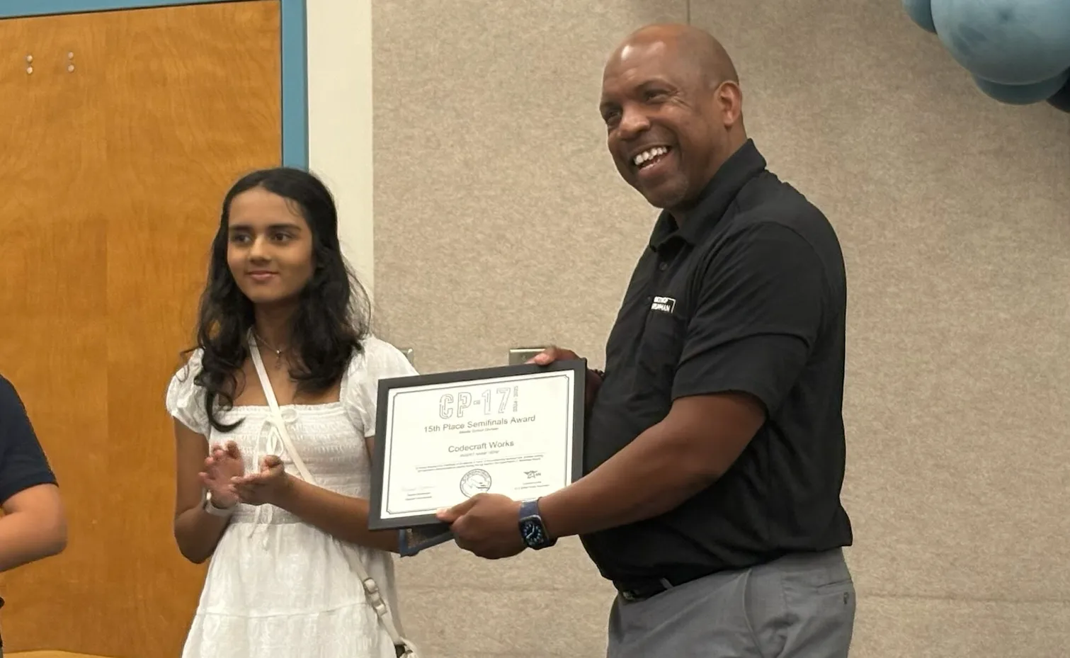 A student wearing a dress clapping her hands, standing next to a smiling man holding a framed certificate. The words "CP-17 Semifinals Award" are visible at the top of the page.