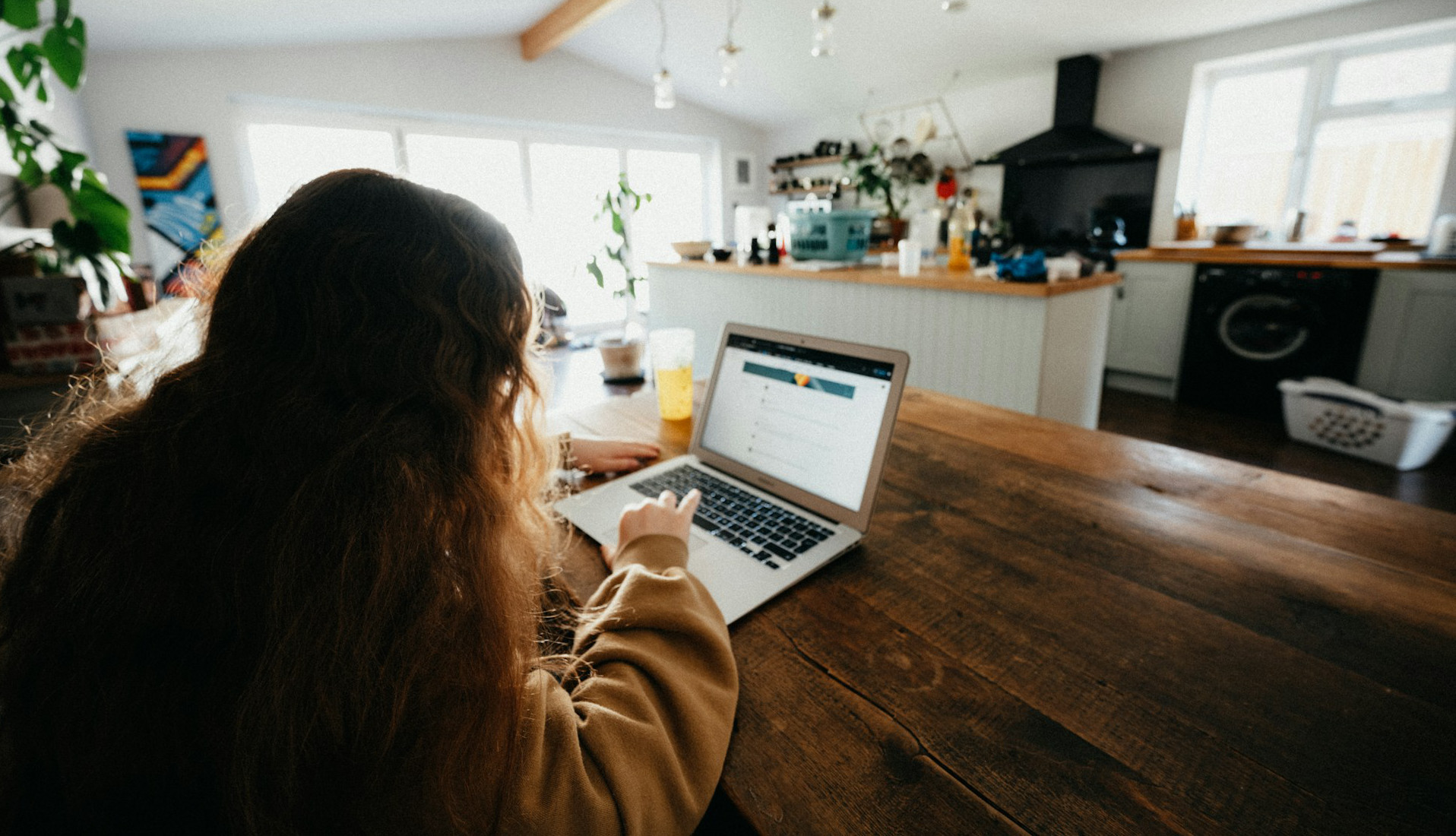 The back of a student with long hear using a silver laptop at their kitchen table, facing their bright, sunlit kitchen